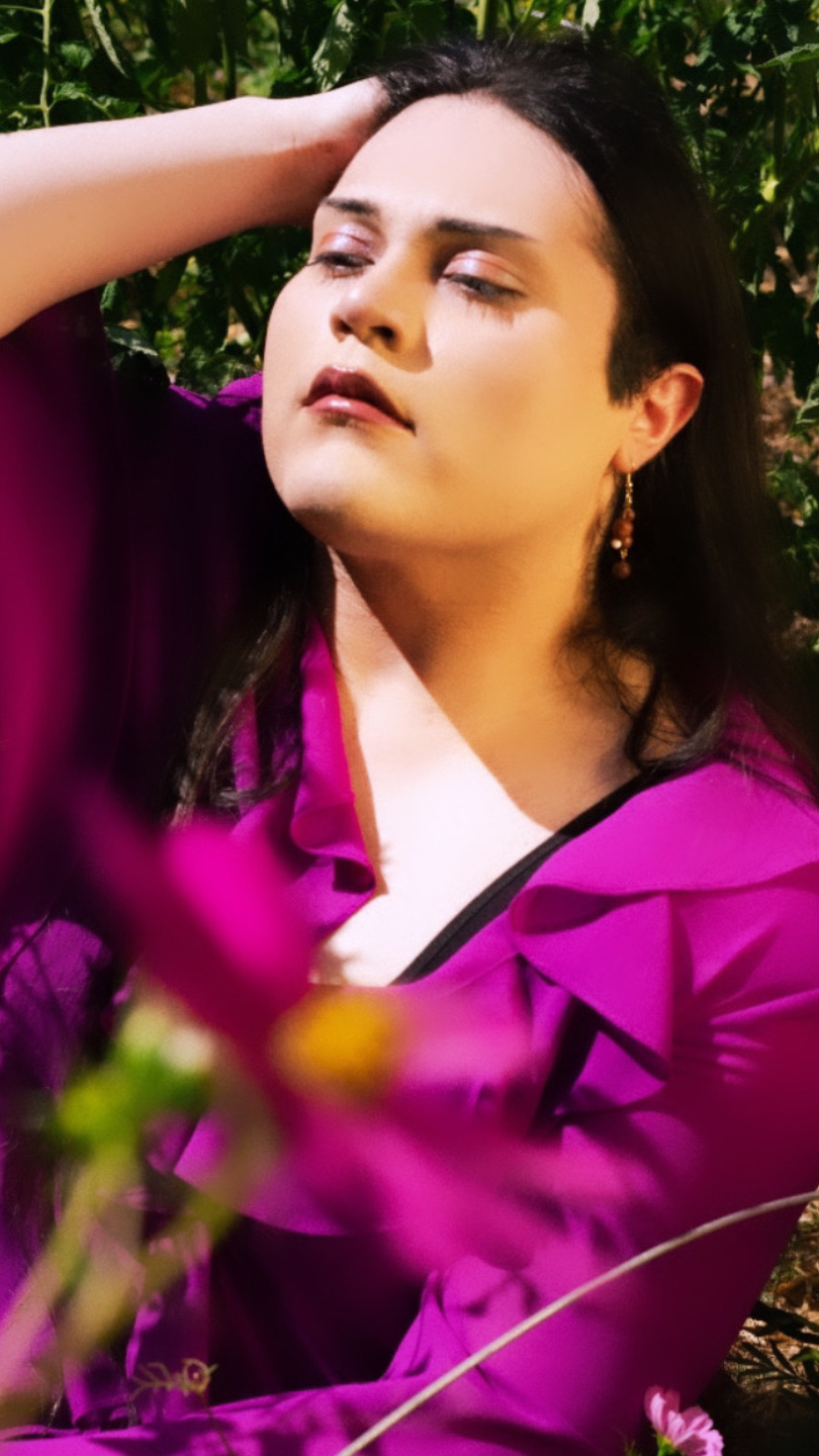 Woman with dark hair and earrings outdoors, wearing a purple jacket and white top, with her hand on her head, eyes closed.