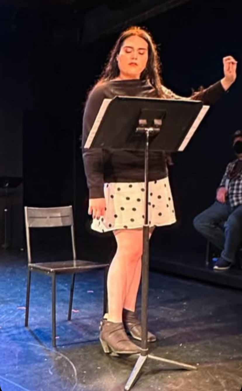 Young woman singing on stage with a music stand, wearing a polka dot skirt and ankle boots, with a chair nearby.