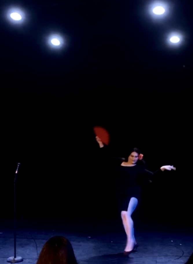 Performer on stage with arms extended, illuminated by stage lights, dark background, person wearing dark clothing and a red hat.