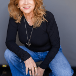 Woman with curly red hair smiling, wearing a black top and blue jeans, crouching against a gray background.
