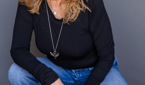 Woman with curly red hair smiling, wearing a black top and blue jeans, crouching against a gray background.