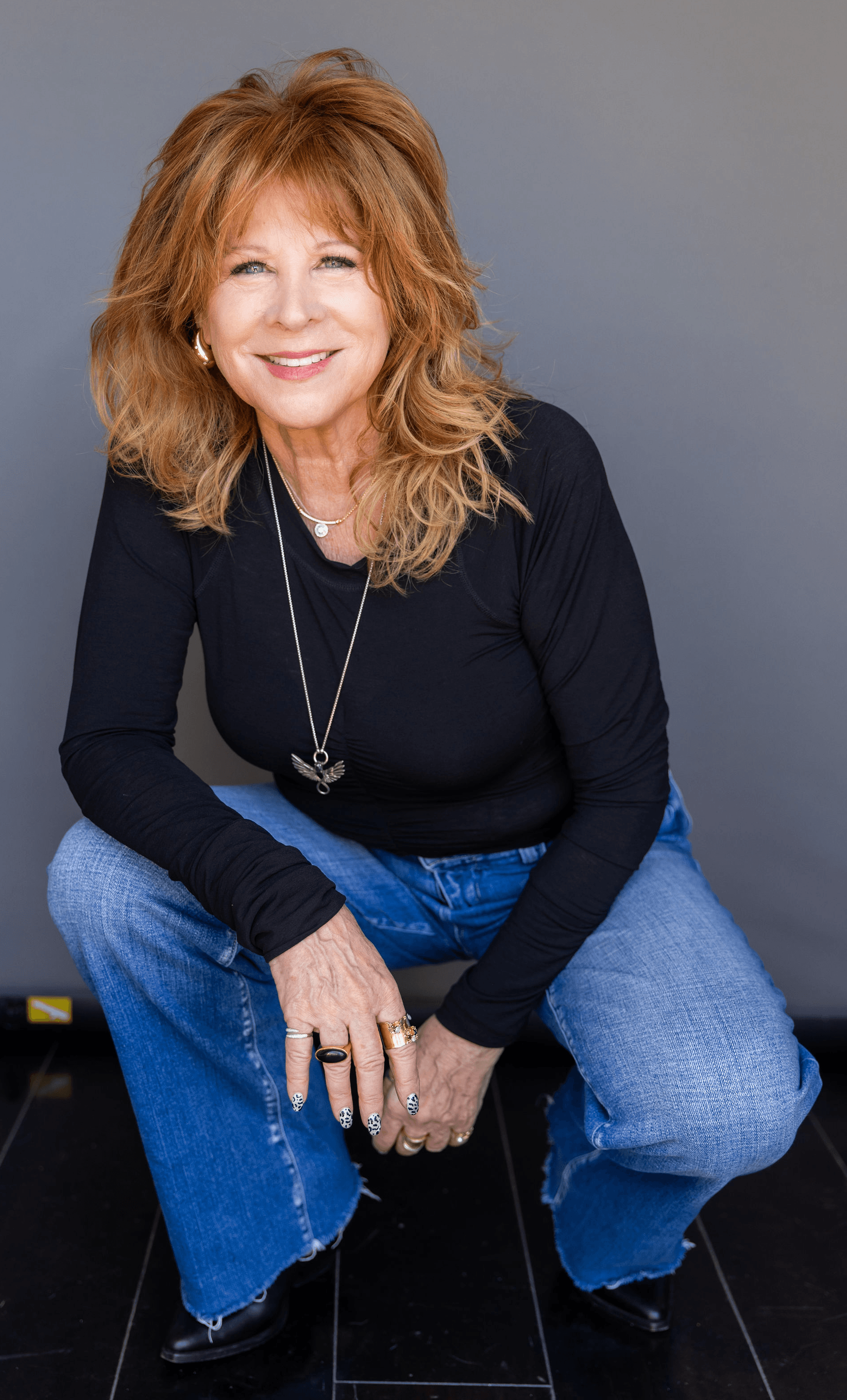 Woman with curly red hair smiling, wearing a black top and blue jeans, crouching against a gray background.