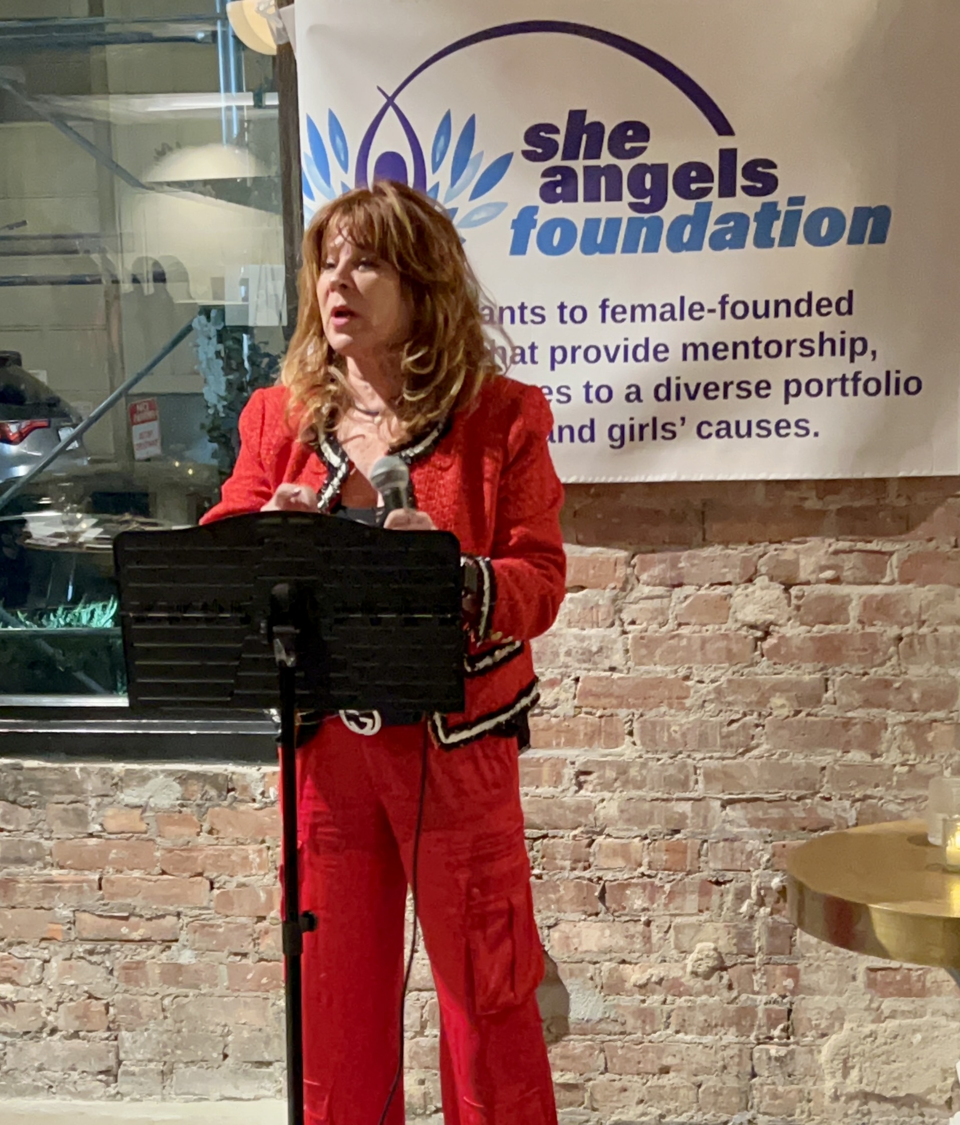 Woman in red suit speaking at a podium with a banner behind her, brick wall background, event setting.