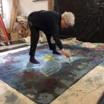 Person with curly white hair painting on large fabric laid on the floor, surrounded by art supplies and boxes.