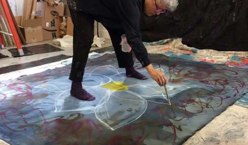 Person with curly white hair painting on large fabric laid on the floor, surrounded by art supplies and boxes.