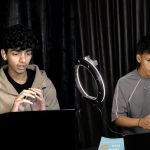 Two young men sit at a table with laptops, in front of black curtains, with ring lights nearby, engaging with devices.