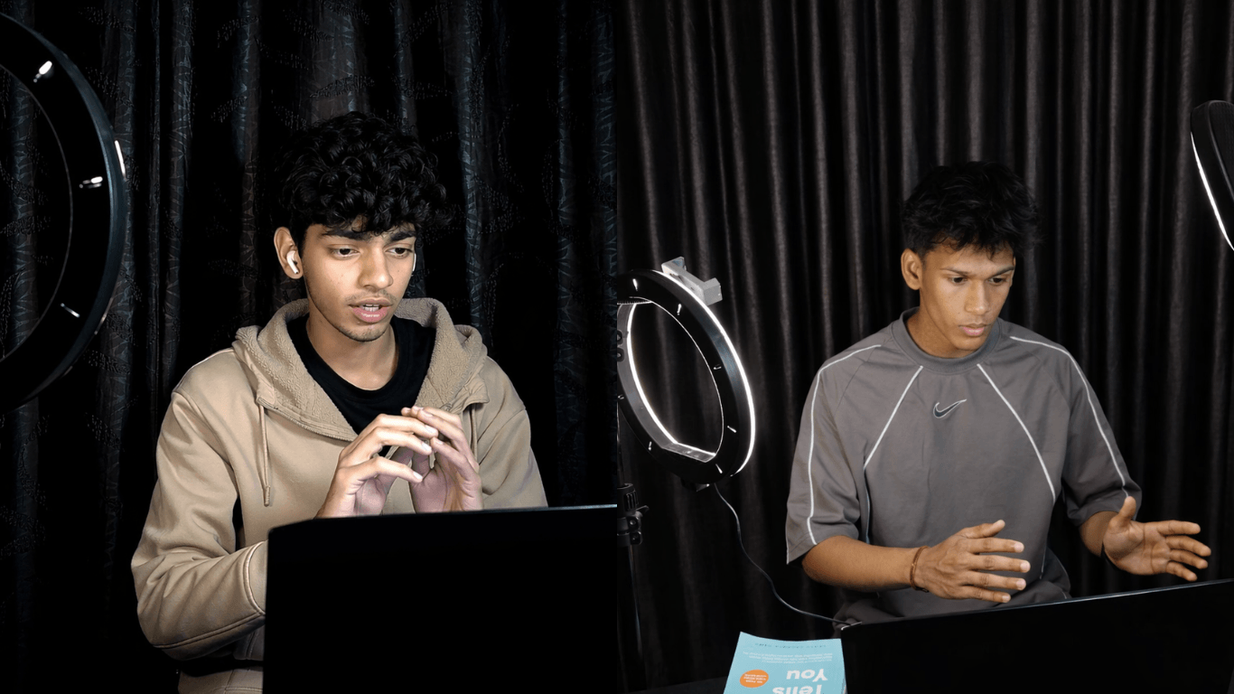 Two young men sit at a table with laptops, in front of black curtains, with ring lights nearby, engaging with devices.