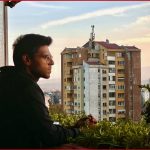 Person sitting at a balcony with a cityscape and mountains in the background during sunset