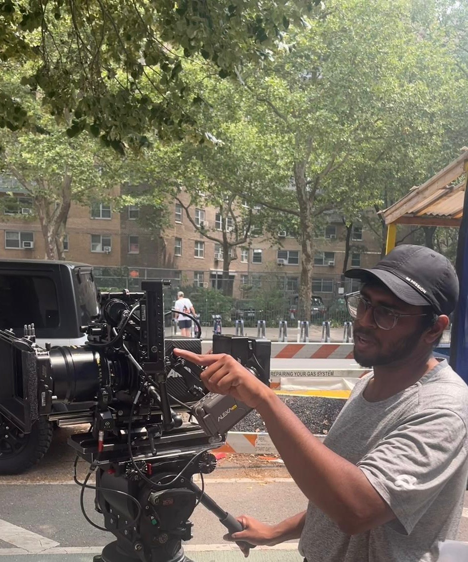 Person operating a professional camera outdoors, trees and buildings in background, wearing a cap and glasses, pointing at the camera.