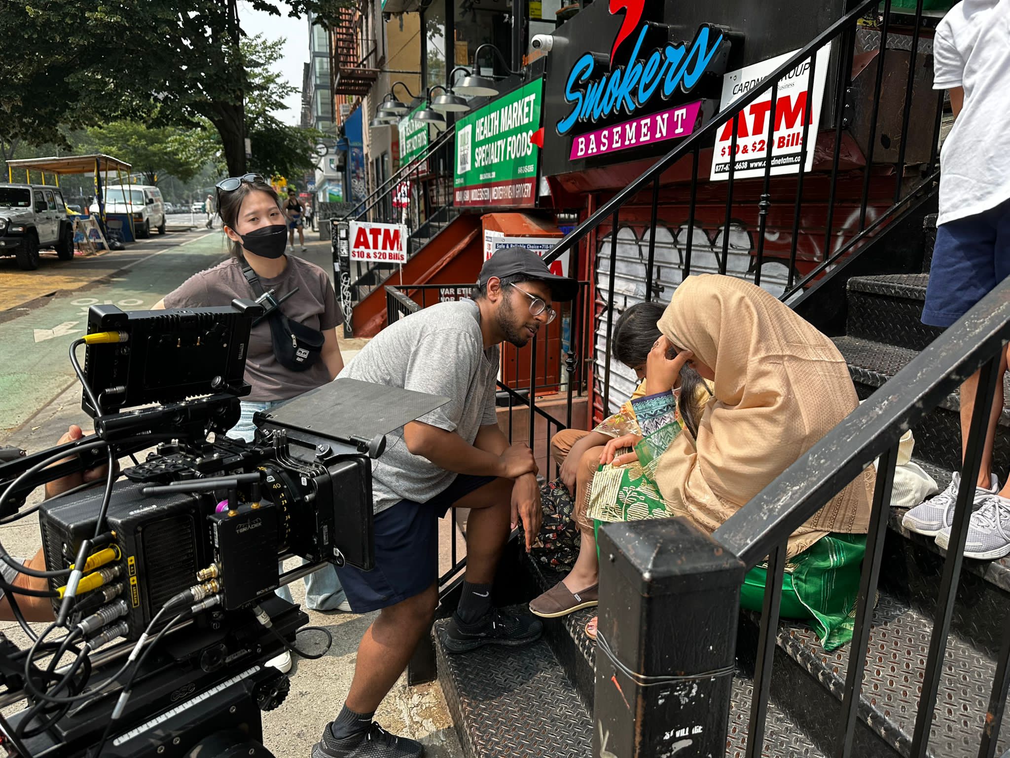 Three people on stairs outside a building, one woman wearing a mask, two men talking, camera equipment nearby.