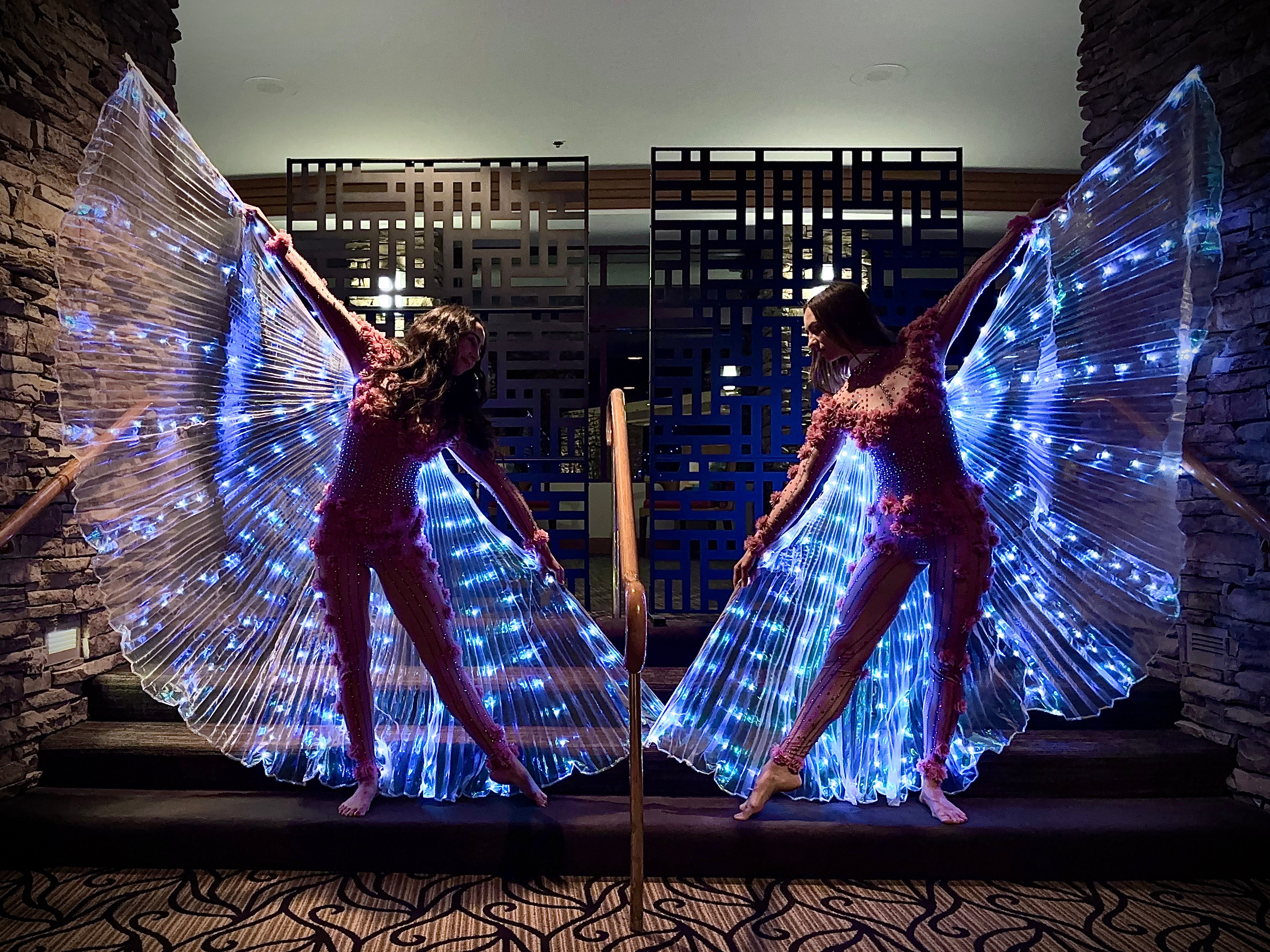 Two performers with illuminated butterfly wings pose indoors, facing each other, with a decorative background and stone wall. Word count: 20.