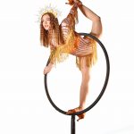 A woman in a tiger-striped costume balances on a circular apparatus, with arms and legs extended, against a white background.