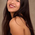 Young woman with long dark hair smiling, wearing a black top, against a neutral background.