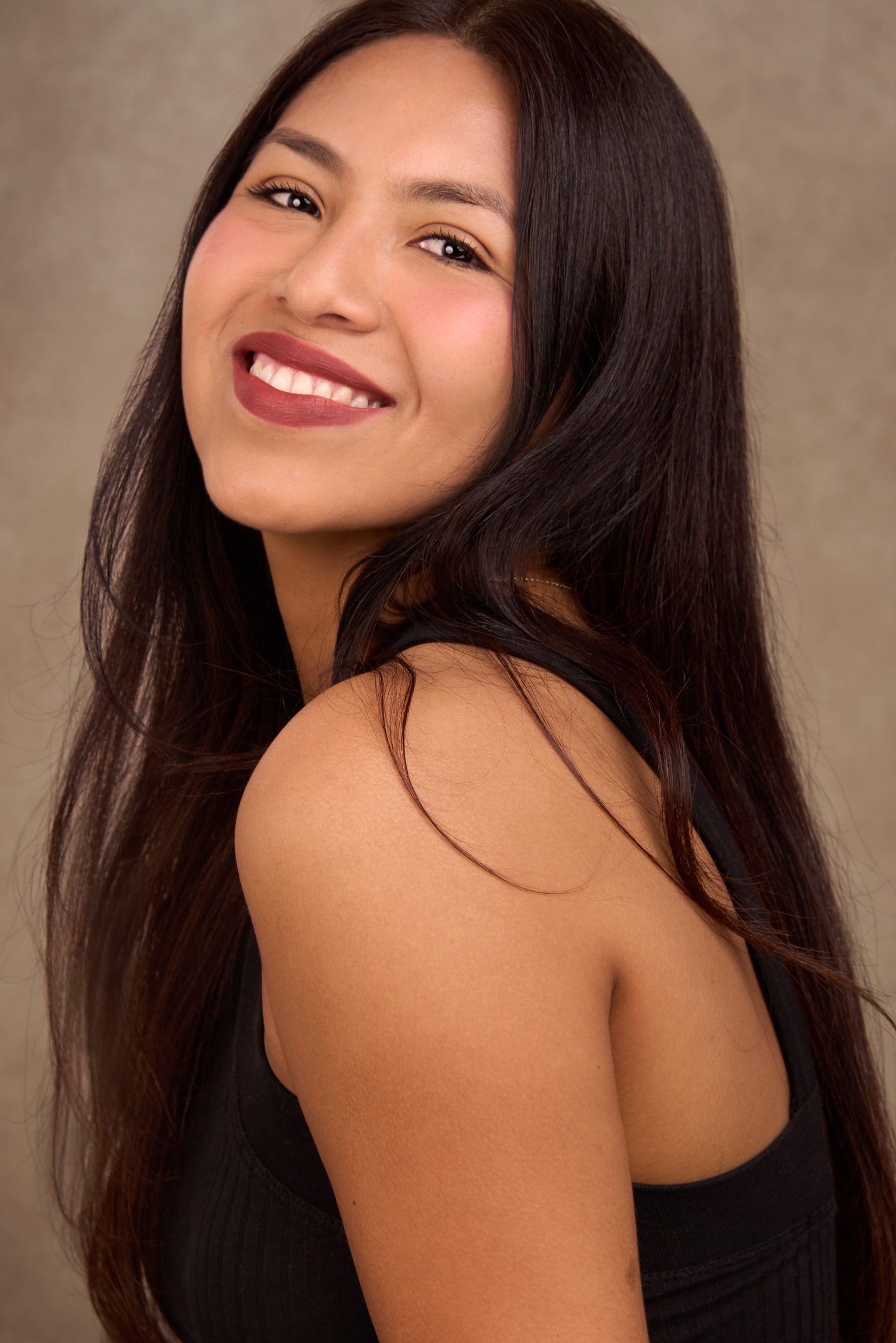 Young woman with long dark hair smiling, wearing a black top, against a neutral background.