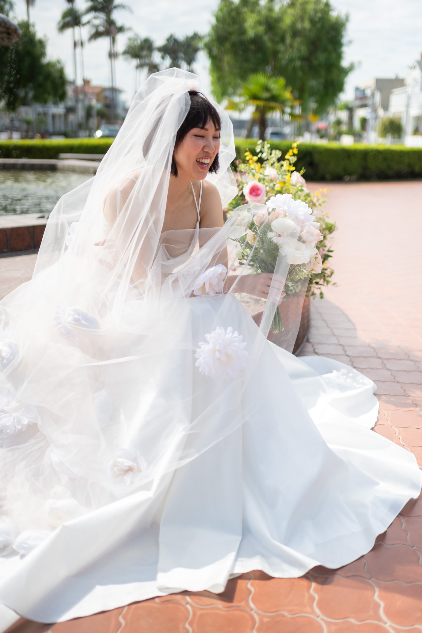 Bride in wedding dress and veil sitting outdoors with a bouquet of flowers, smiling, on a sunny day.