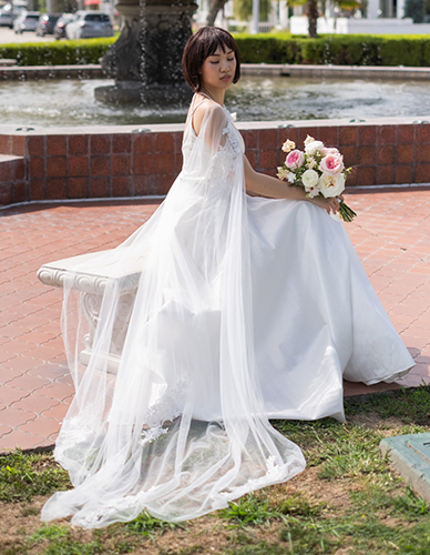 Young woman in a white dress sitting outdoors, holding a bouquet of pink and white flowers, near a fountain.