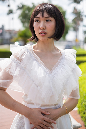 Young woman with short black hair wearing a white ruffled blouse outdoors, with trees and sky in background.
