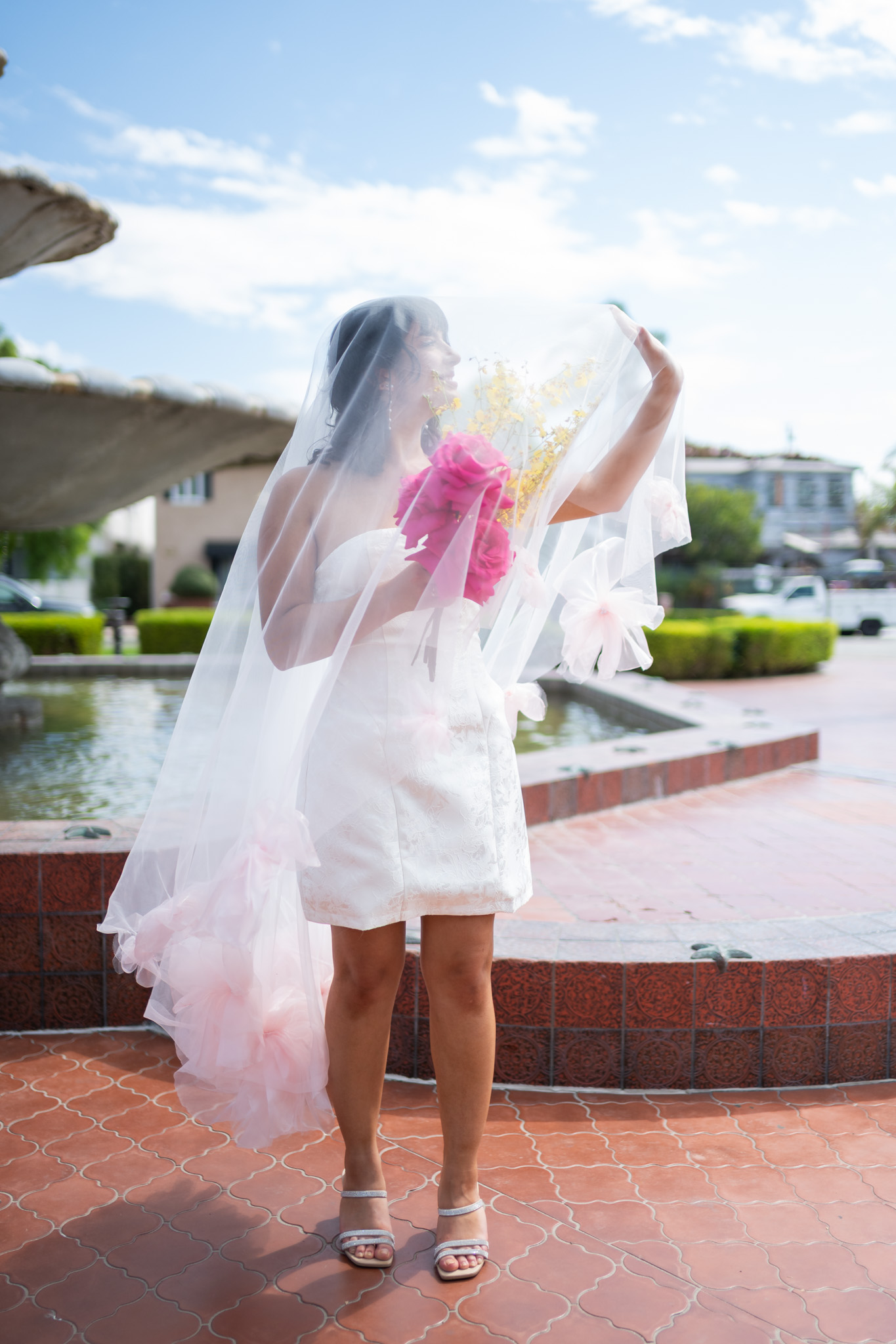 Person in a white dress and veil holding pink flowers, standing outdoors near a fountain, with a blue sky and buildings in background.