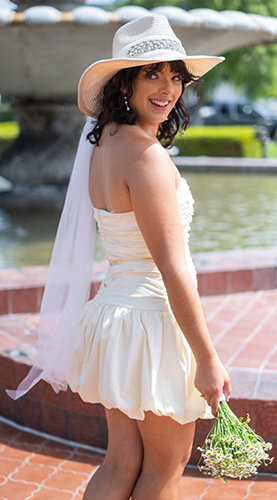 Woman in a white dress and wide-brimmed hat holding a small bouquet, smiling outdoors near a fountain.