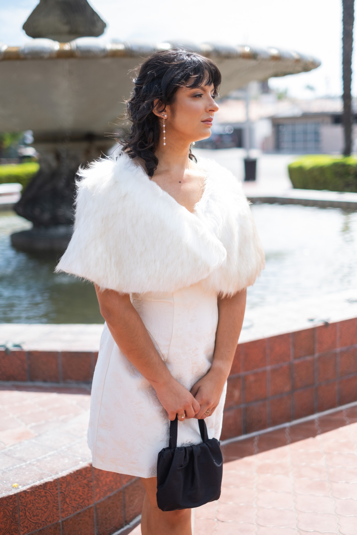 Woman in white dress and fur stole holding a black handbag near a fountain outdoors.