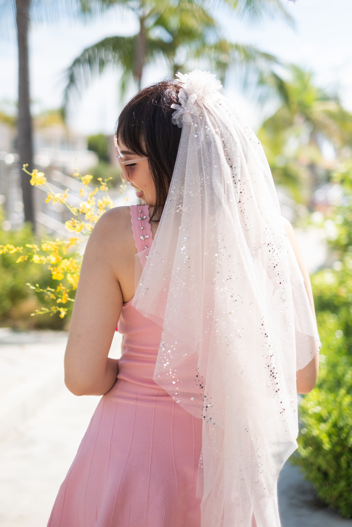 Young woman in pink dress with veil outdoors, surrounded by greenery and yellow flowers, facing sideways with head slightly down.