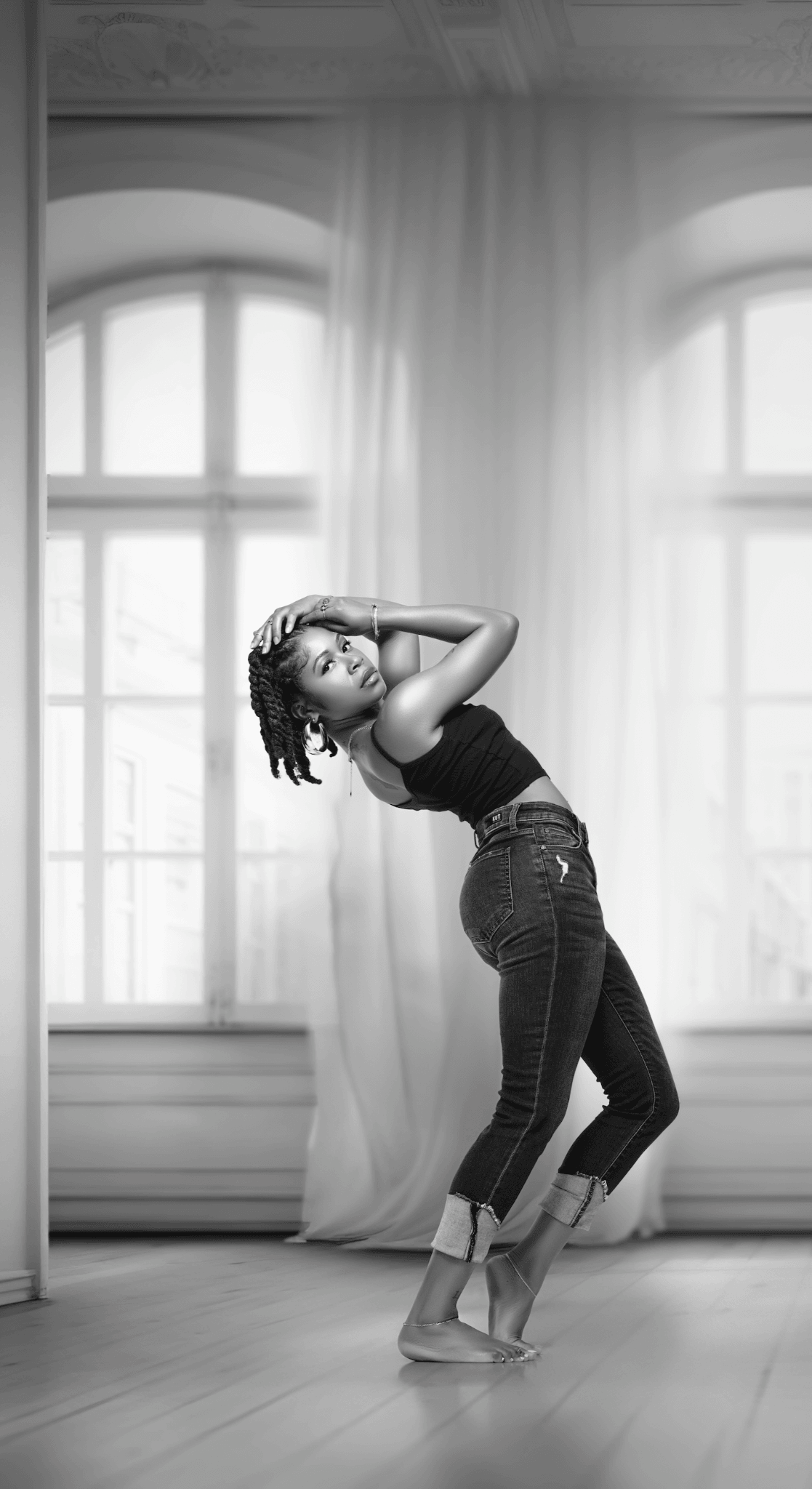 Young woman with dreadlocks poses in front of large windows with curtains, standing on a wooden floor, arms raised behind her head.