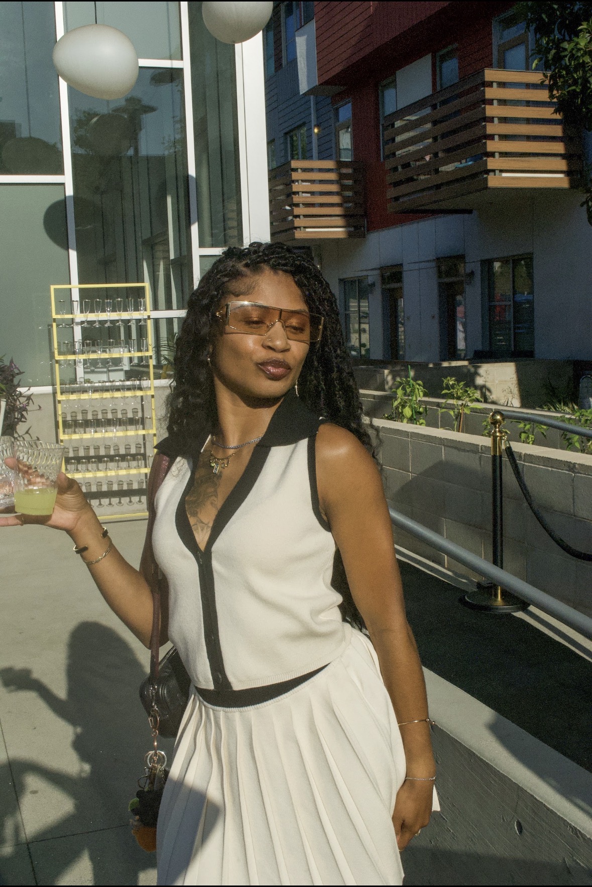 Woman with curly hair and sunglasses standing outdoors in sunlight, wearing a sleeveless white dress with black accents.