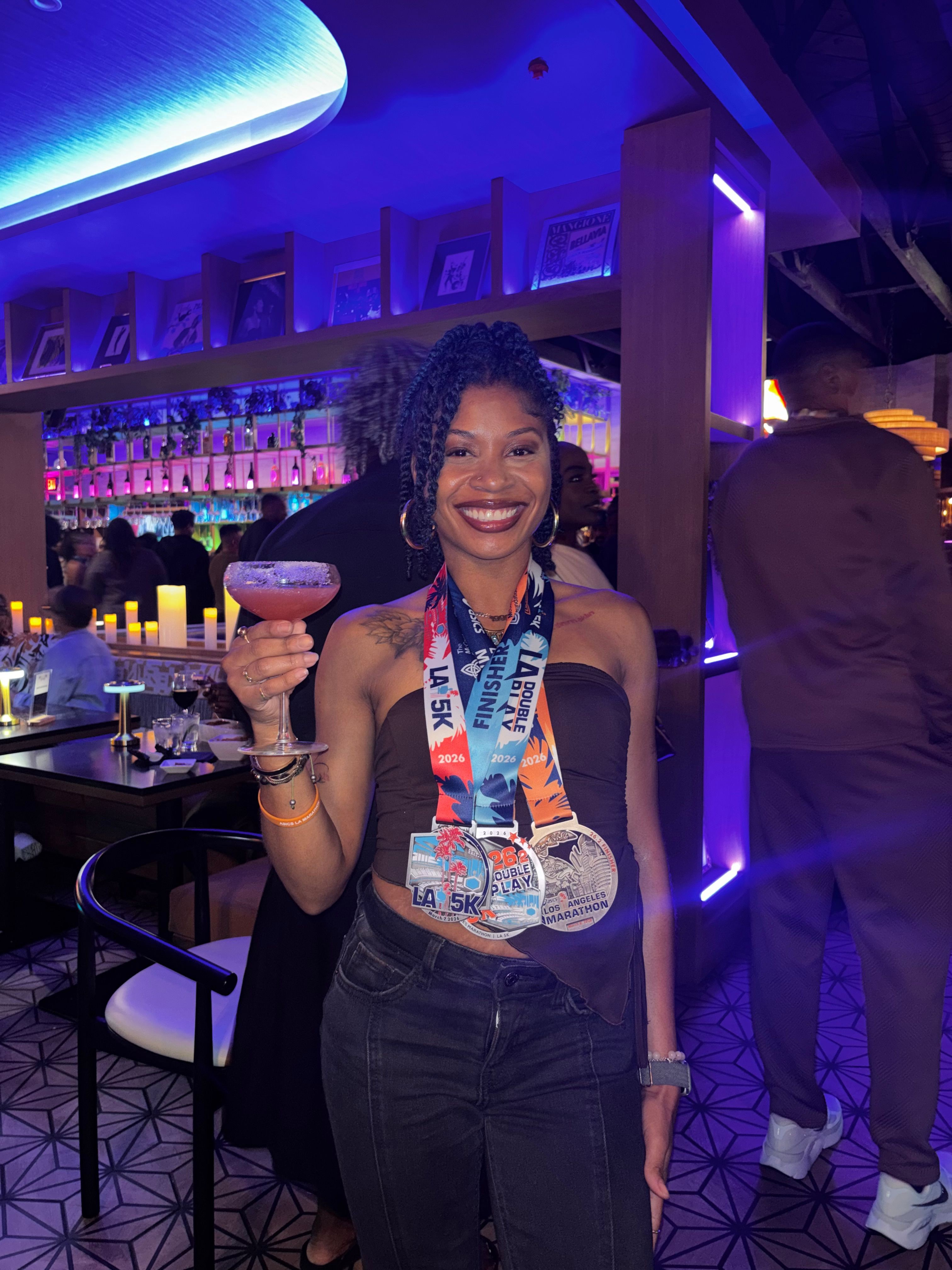 Woman with curly hair smiling, holding a drink, in a bar with purple lighting and shelves of bottles.