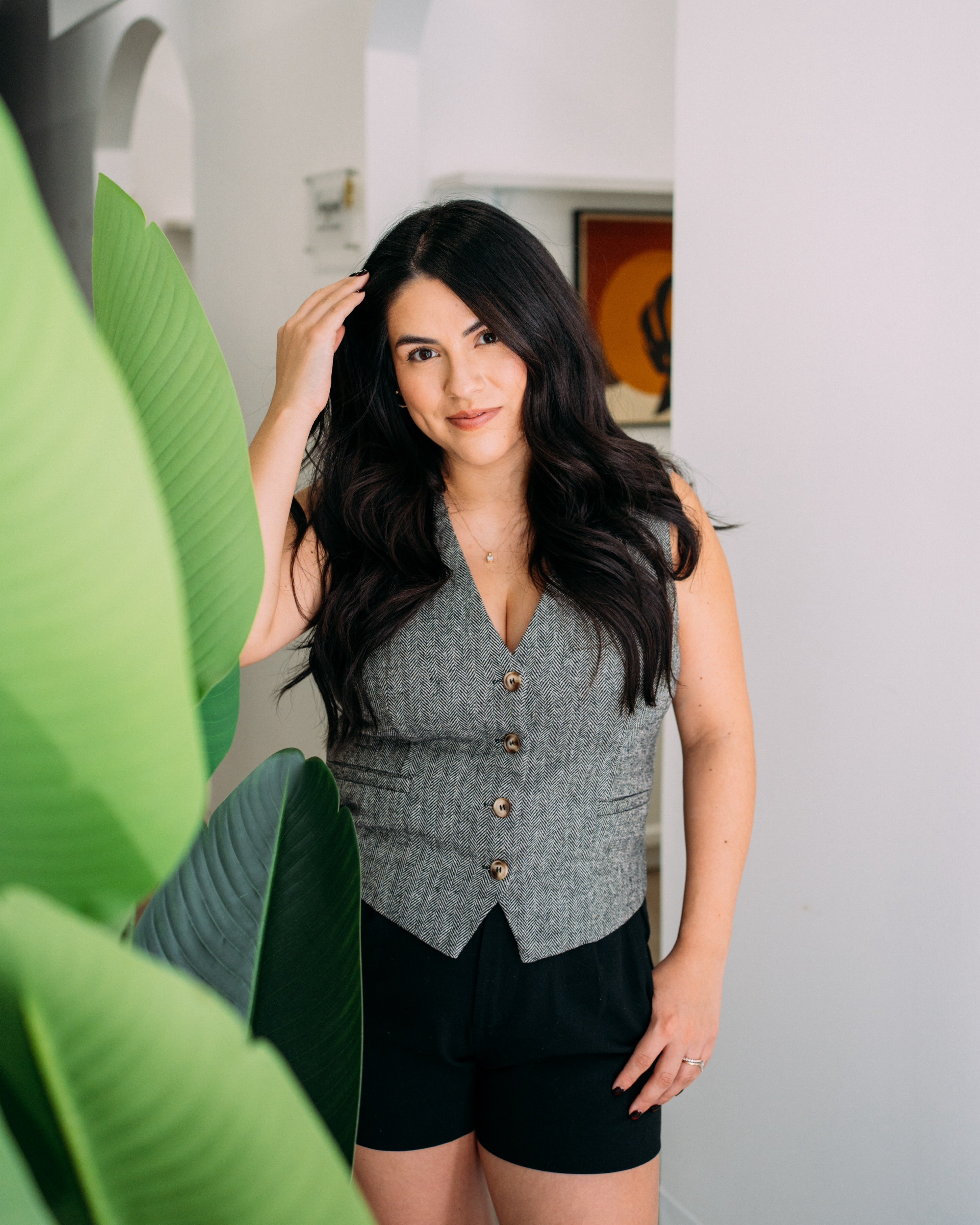 Young woman with long dark hair, wearing a gray vest and black shorts, standing indoors near a white wall and green plant.