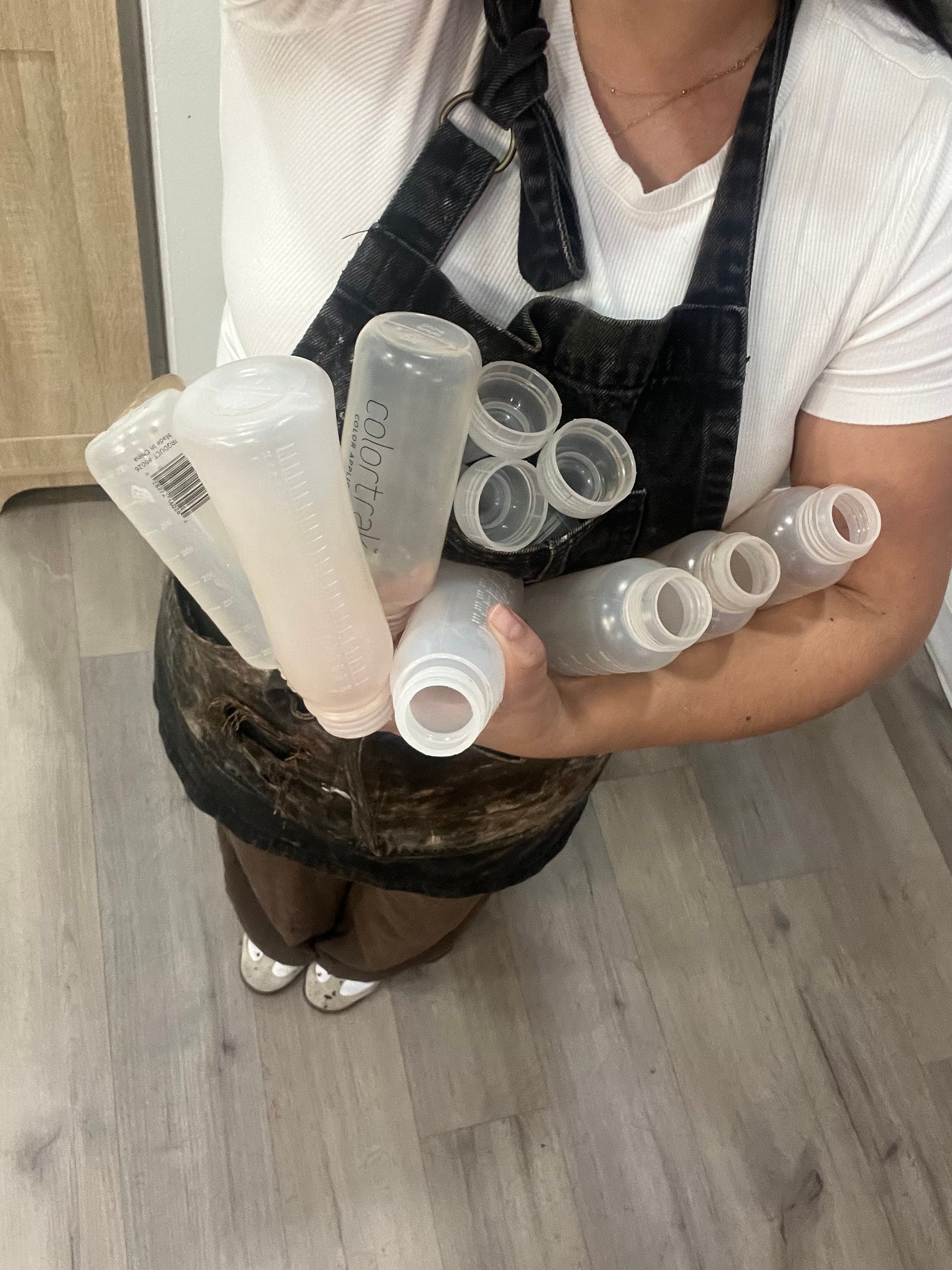 Person holding multiple empty plastic bottles, standing on a wooden floor, wearing a white shirt and dark pants.