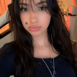 Young woman with long dark hair, black flower hair accessory, star face tattoo, wearing a dark top, indoors.