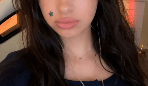 Young woman with long dark hair, black flower hair accessory, star face tattoo, wearing a dark top, indoors.