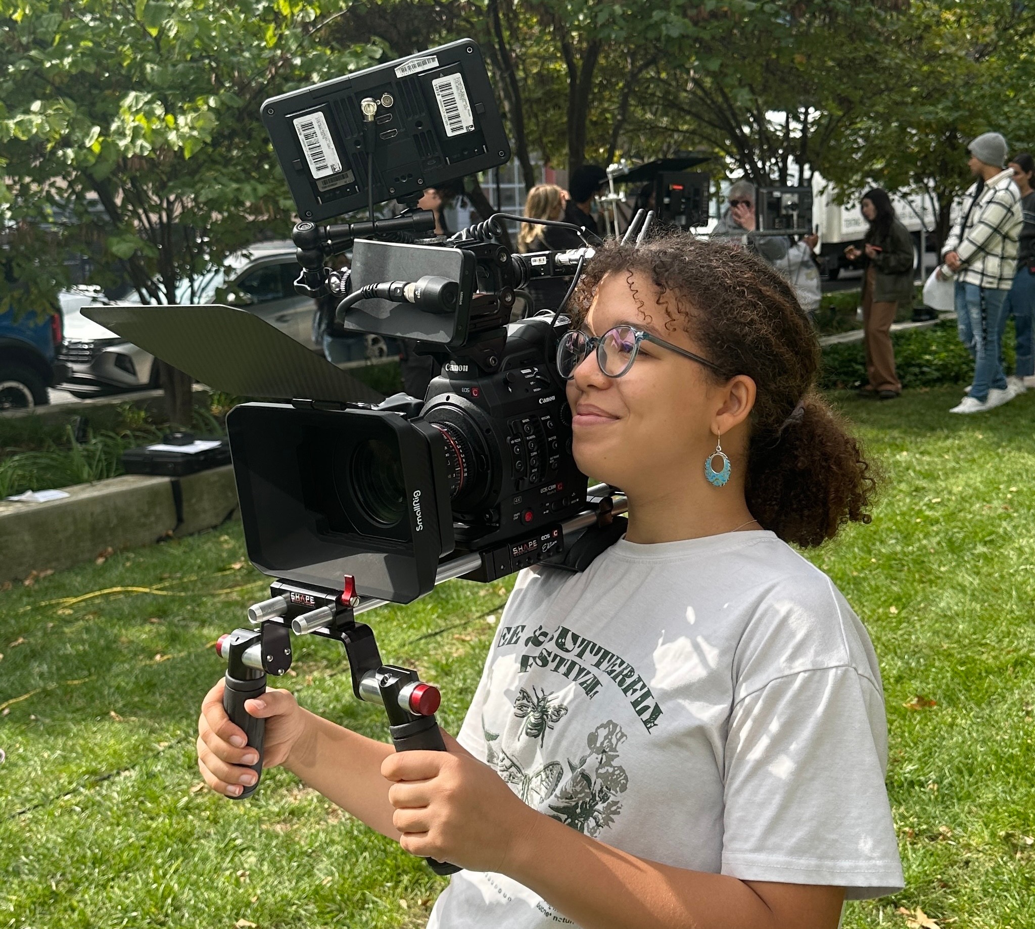 Young woman operating a professional video camera outdoors on grass, with trees and people in the background.