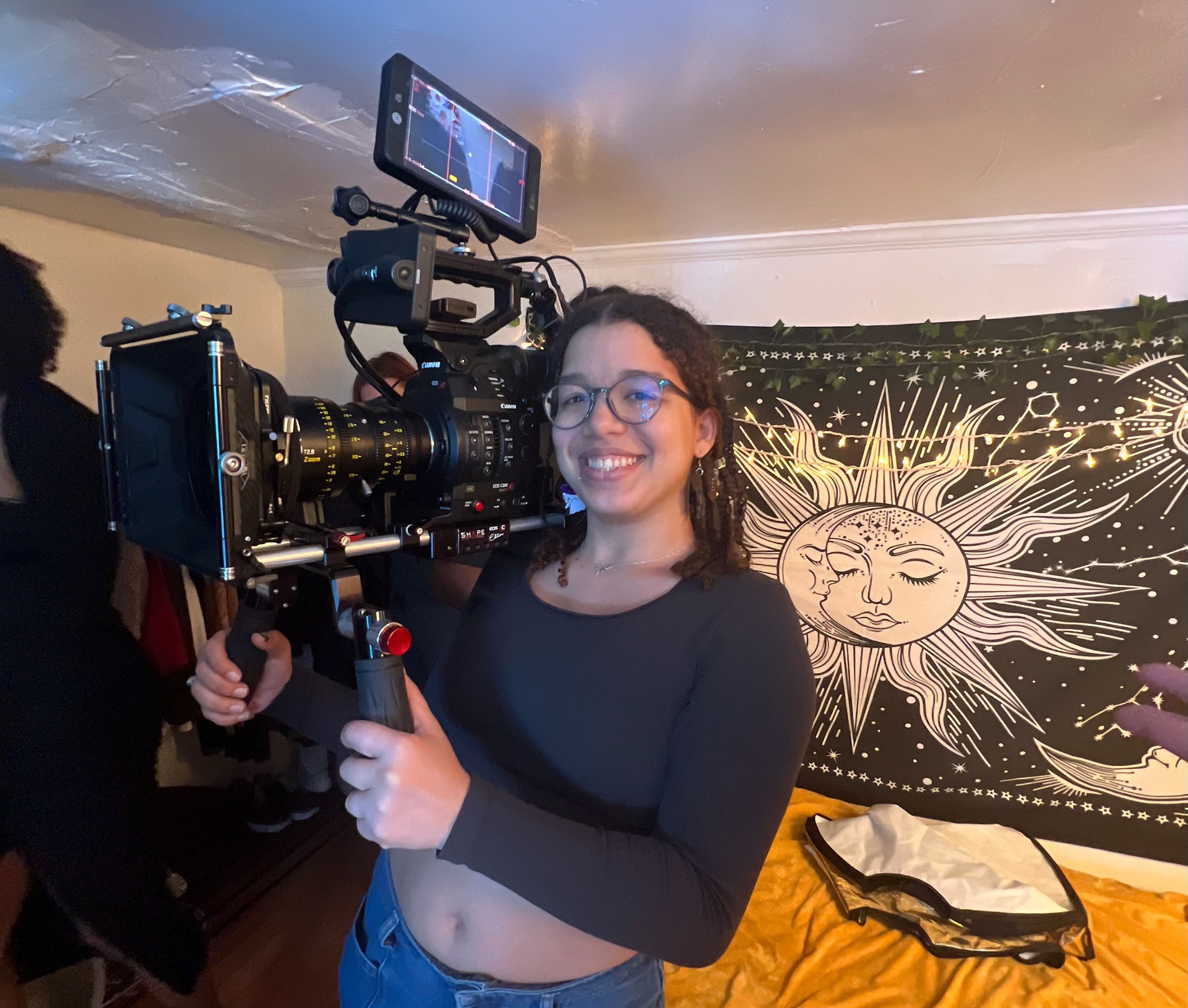 Young woman with glasses holding a camera rig, smiling indoors with a sun and moon tapestry on the wall.