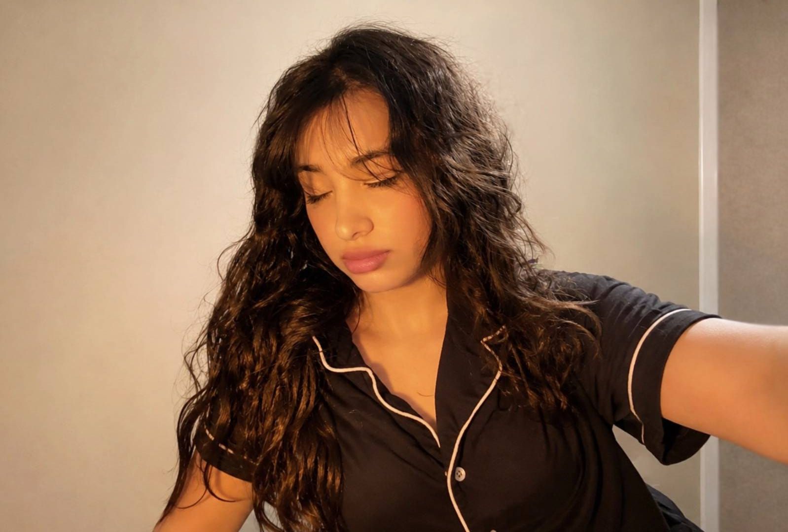 Woman with long, curly hair lying down with eyes closed, wearing a black shirt with white trim, against a plain wall.