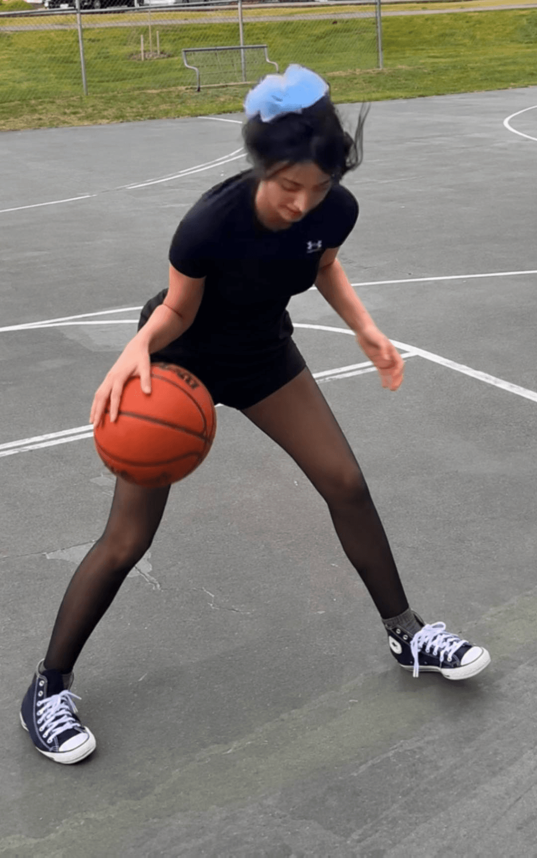 Person playing basketball on outdoor court, wearing black outfit, blue cap, and sneakers, holding basketball with one hand.