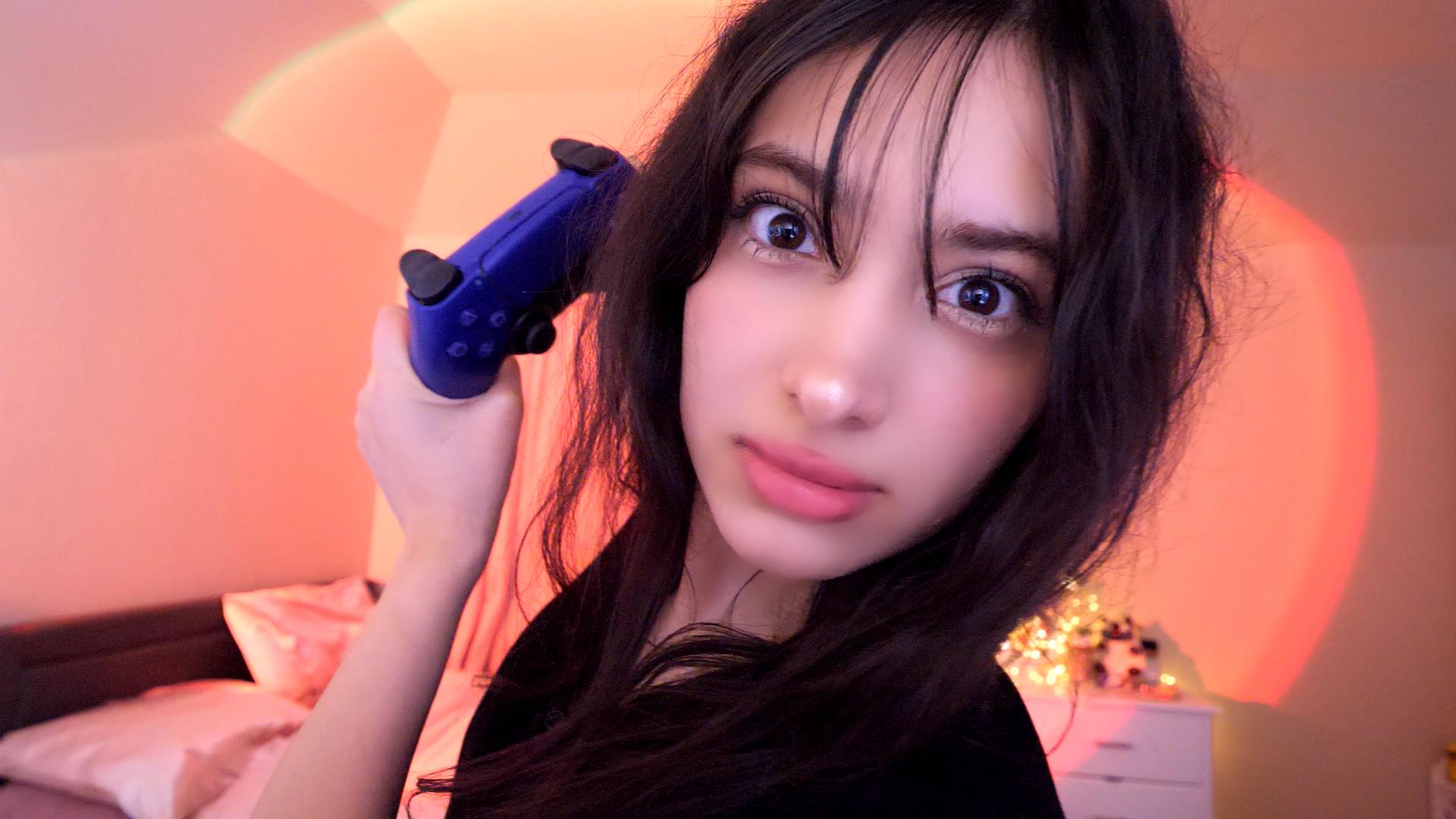 Young woman with long dark hair holding a blue object near her head, in a warmly lit room with a bed and nightstand.