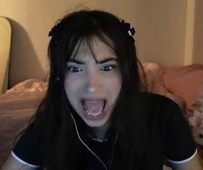 Young woman with dark hair and bangs, wearing a black shirt, appears surprised or shocked, in a bedroom setting.