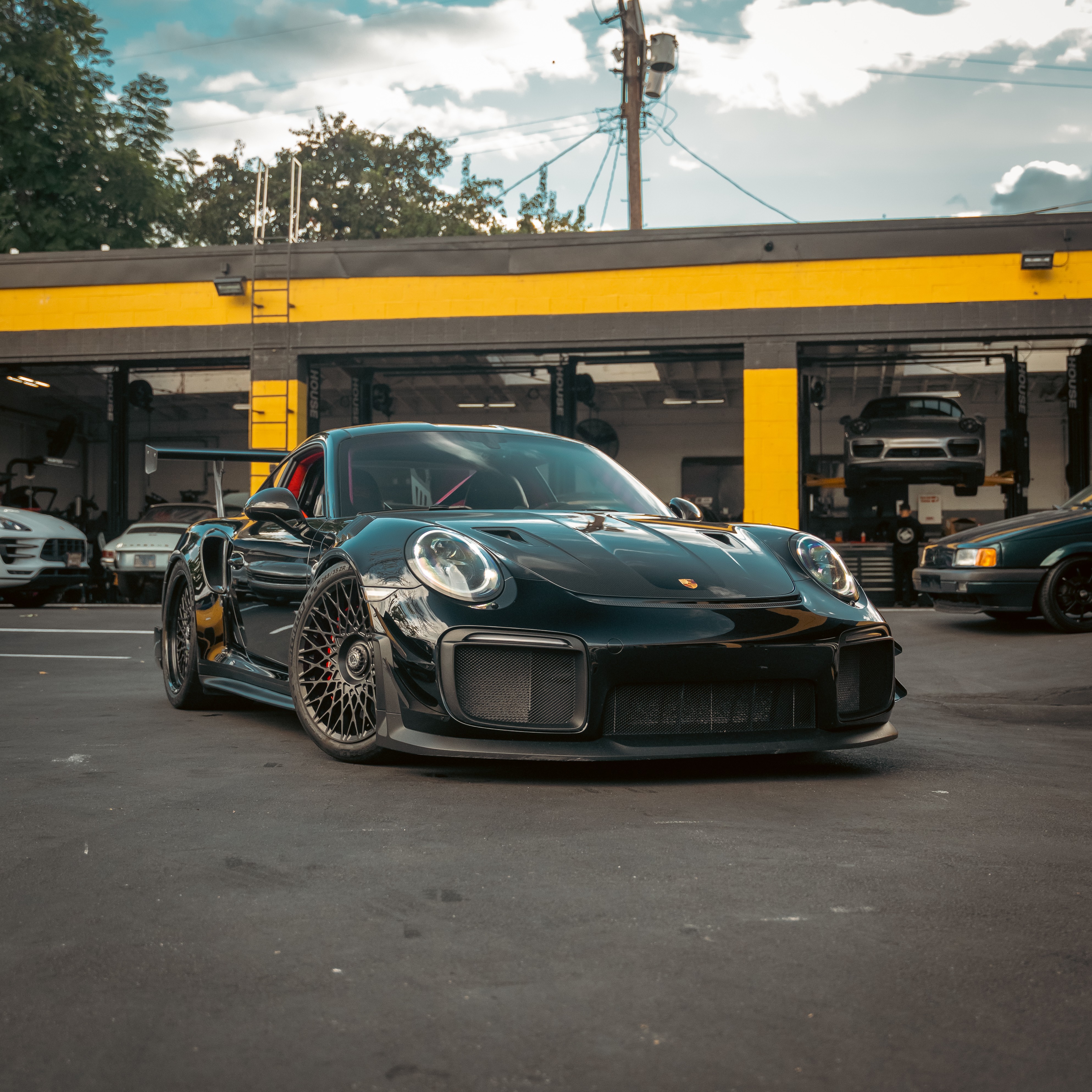 Black sports car parked in front of a yellow and black building with other vehicles in the background, under a partly cloudy sky.