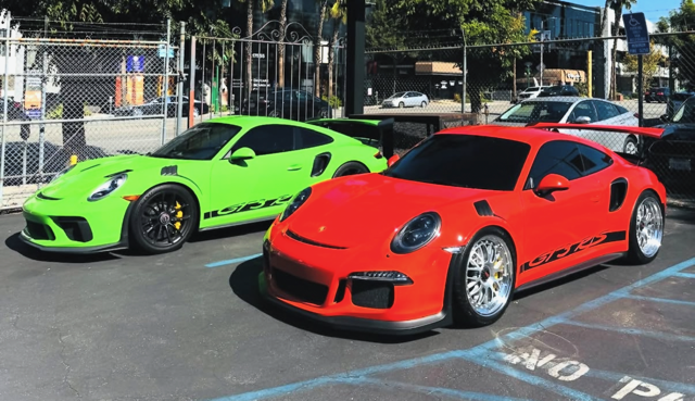 Two sports cars, one green and one red, parked side by side in a parking lot with a fence and trees in background.