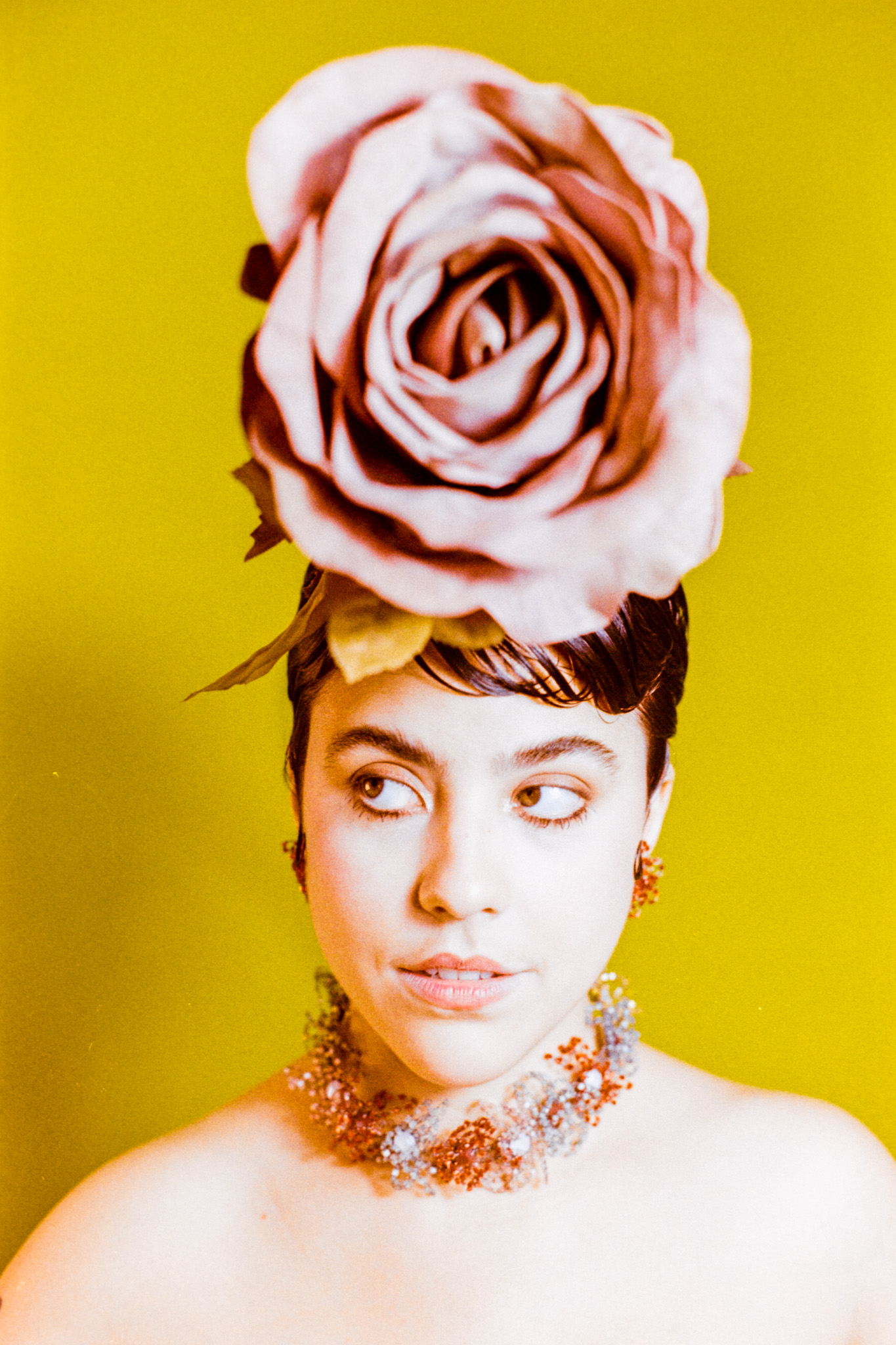 Woman with large pink rose headpiece, wearing a necklace, against yellow background.