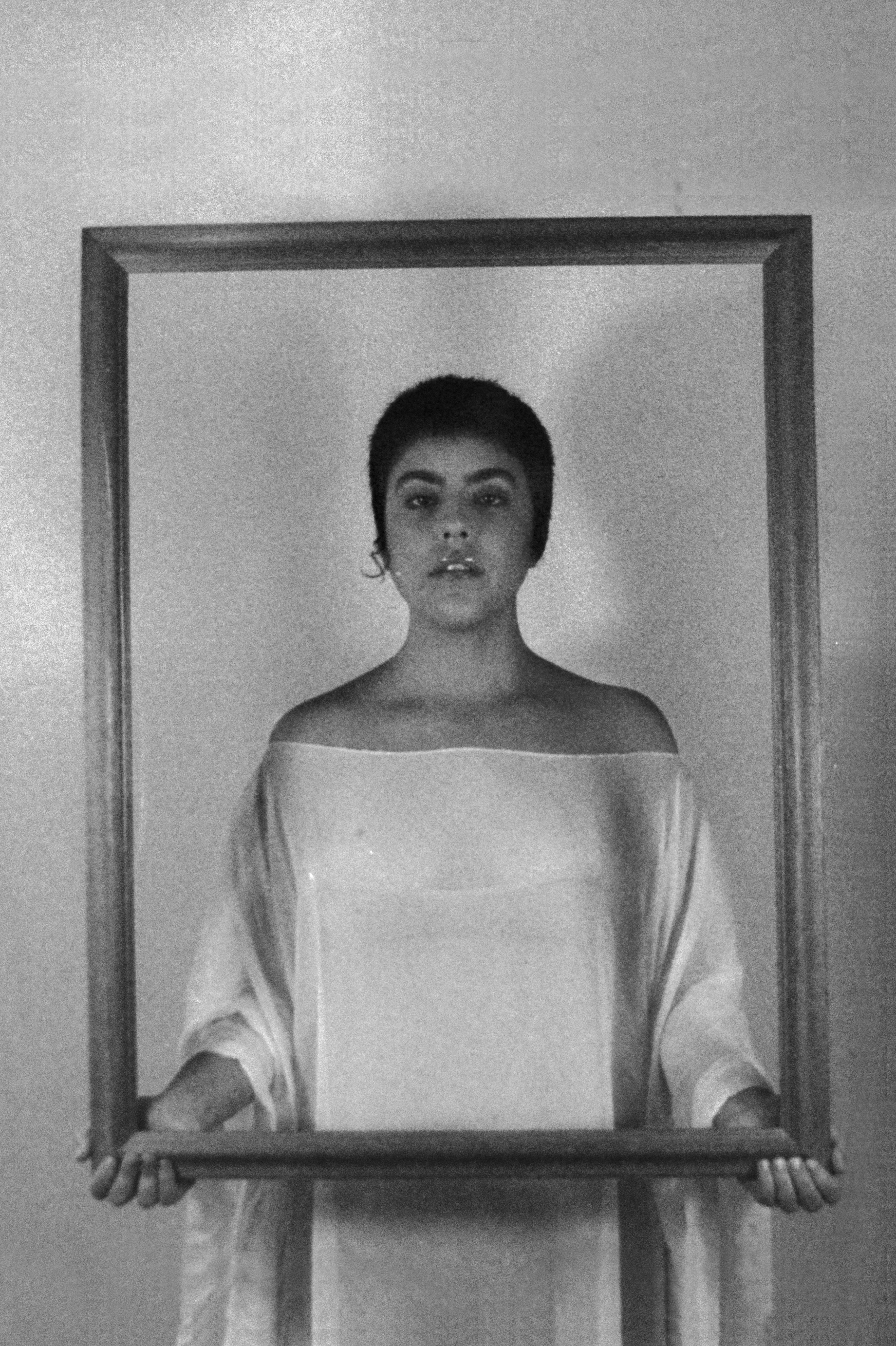 Person holding a frame, looking directly at the camera, with short hair and an off-the-shoulder top, in black and white.