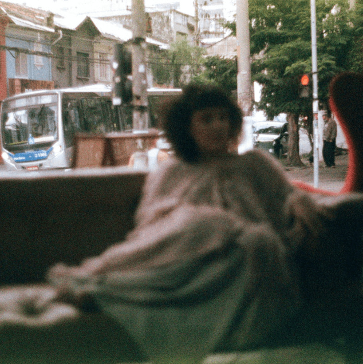 Blurry person with curly hair sitting indoors near a window, city street visible outside, with vehicles and buildings.