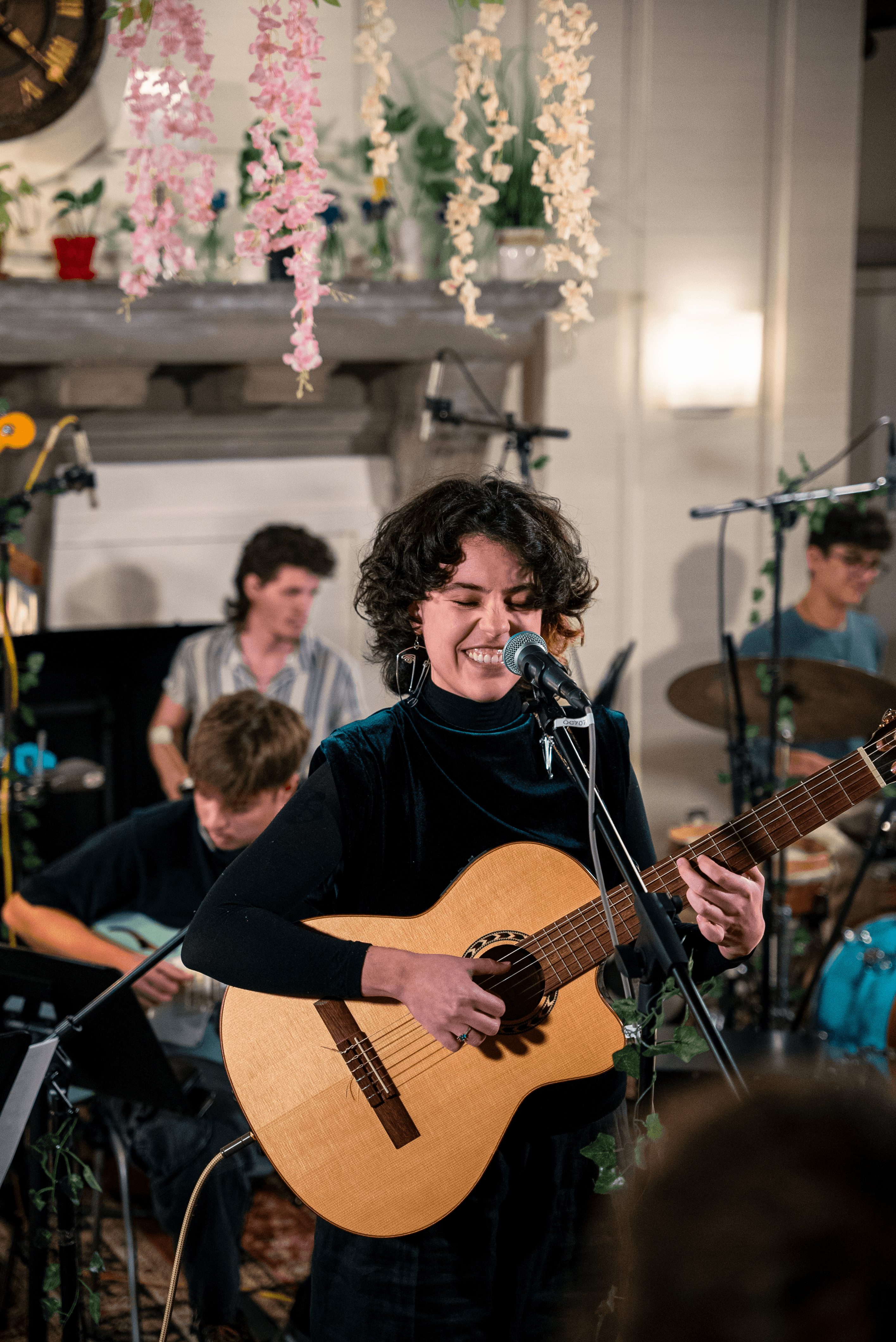 Woman playing guitar and singing with a band in a decorated indoor setting.