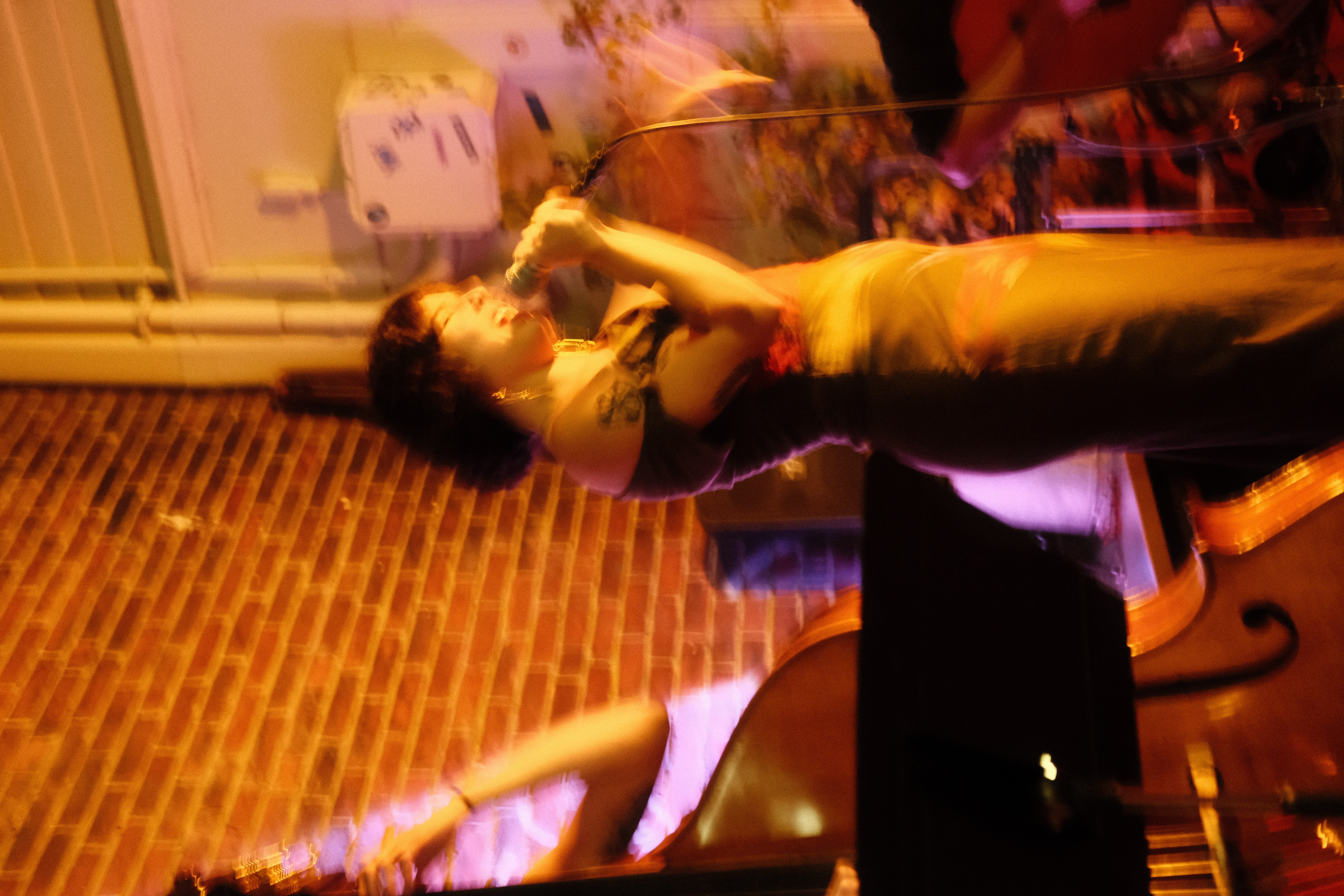 Person singing into a microphone on stage with a brick wall background, stage lighting, and a music stand nearby.