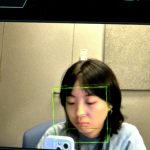 Young woman with dark hair looking down, reflected in a mirror with a green face detection box, in a room with beige walls