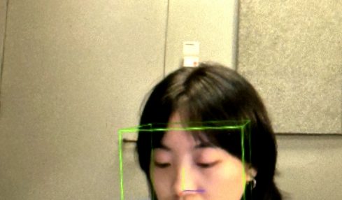 Young woman with dark hair looking down, reflected in a mirror with a green face detection box, in a room with beige walls