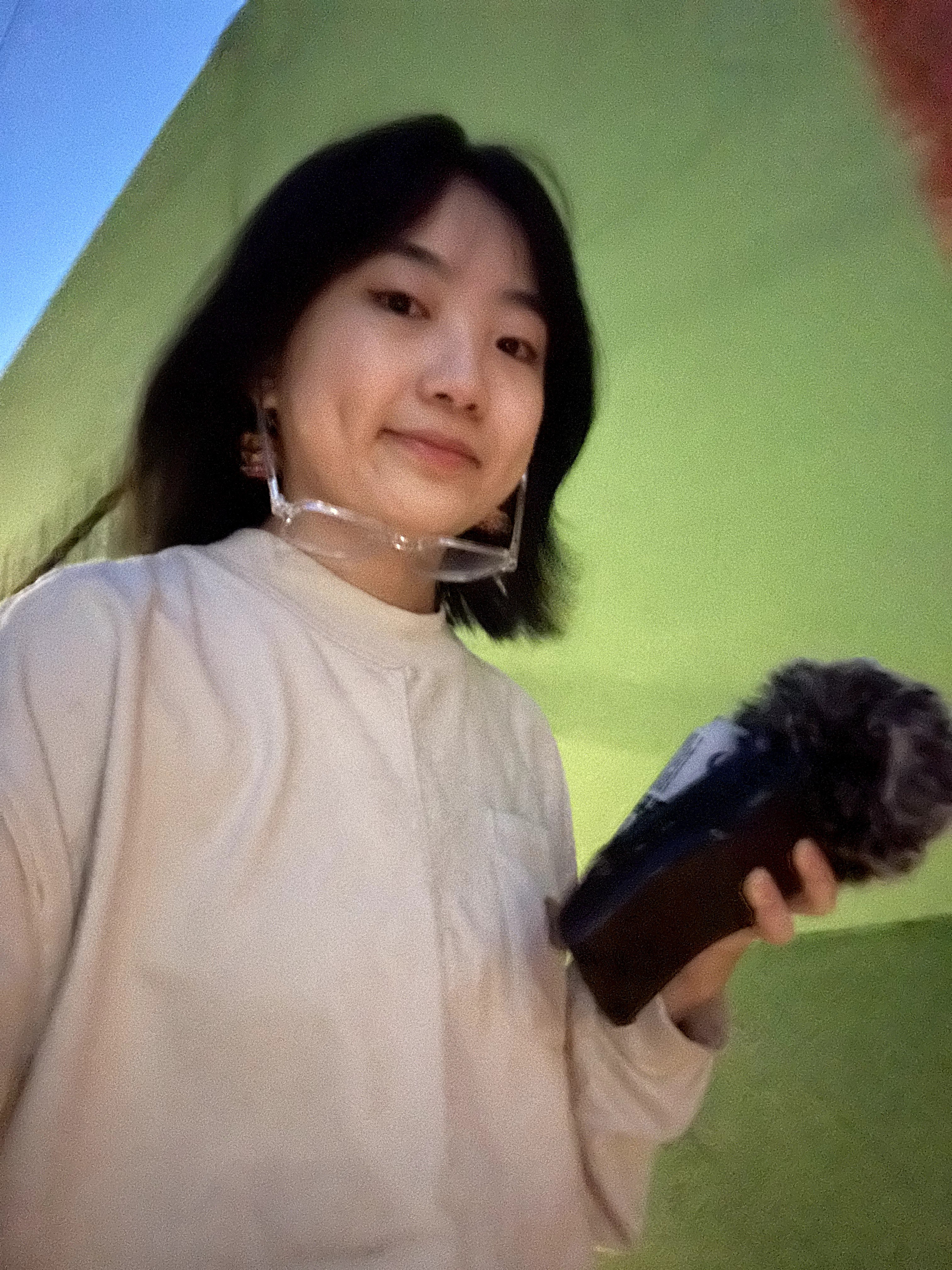 Person with shoulder-length dark hair holding a microphone, standing in front of a green background.