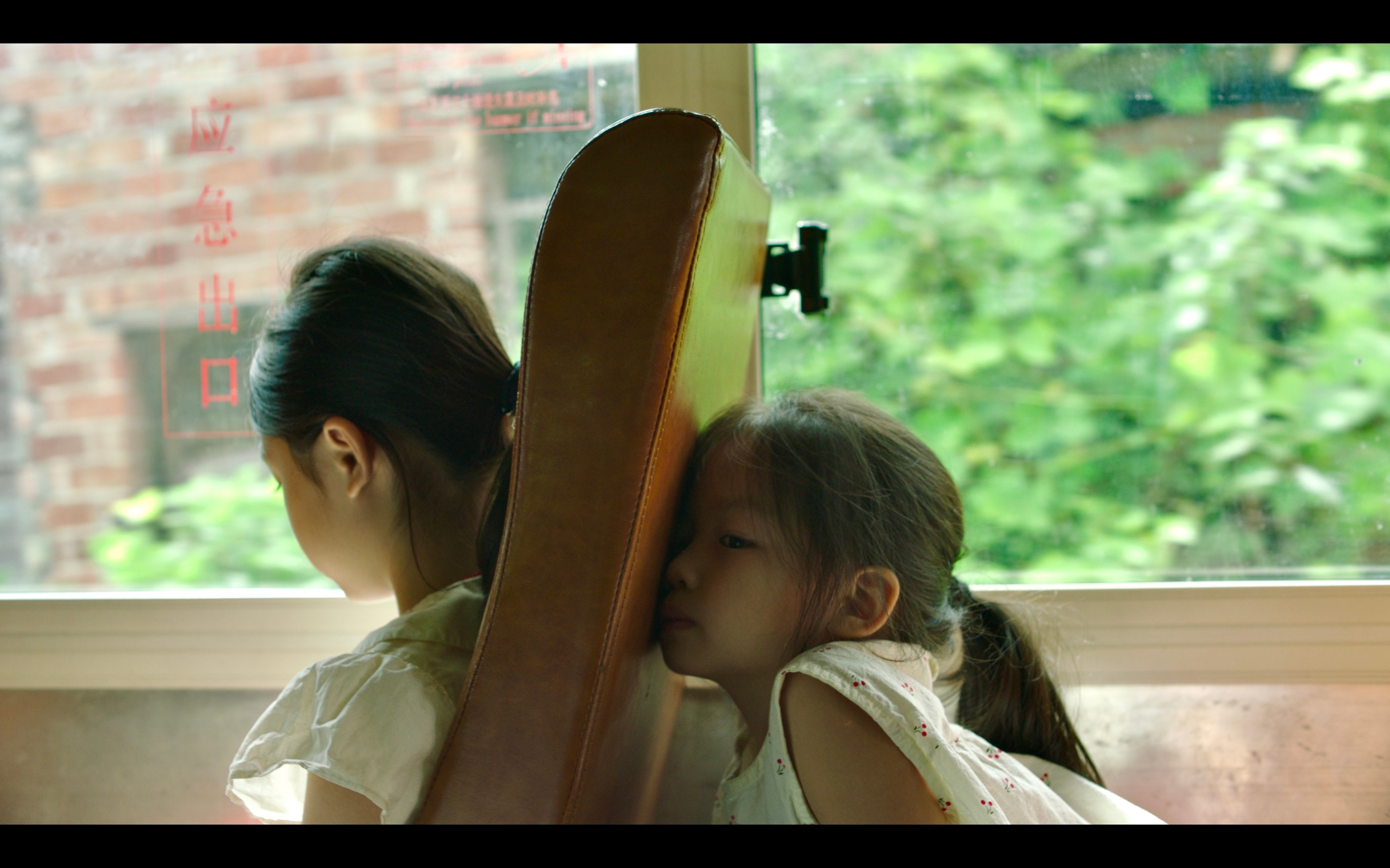 Two young girls sit by a window, one leaning on a skateboard, looking outside with greenery visible through the window.