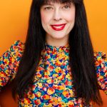 Woman with long dark hair, red lipstick, smiling, wearing a colorful patterned top, standing against an orange background.